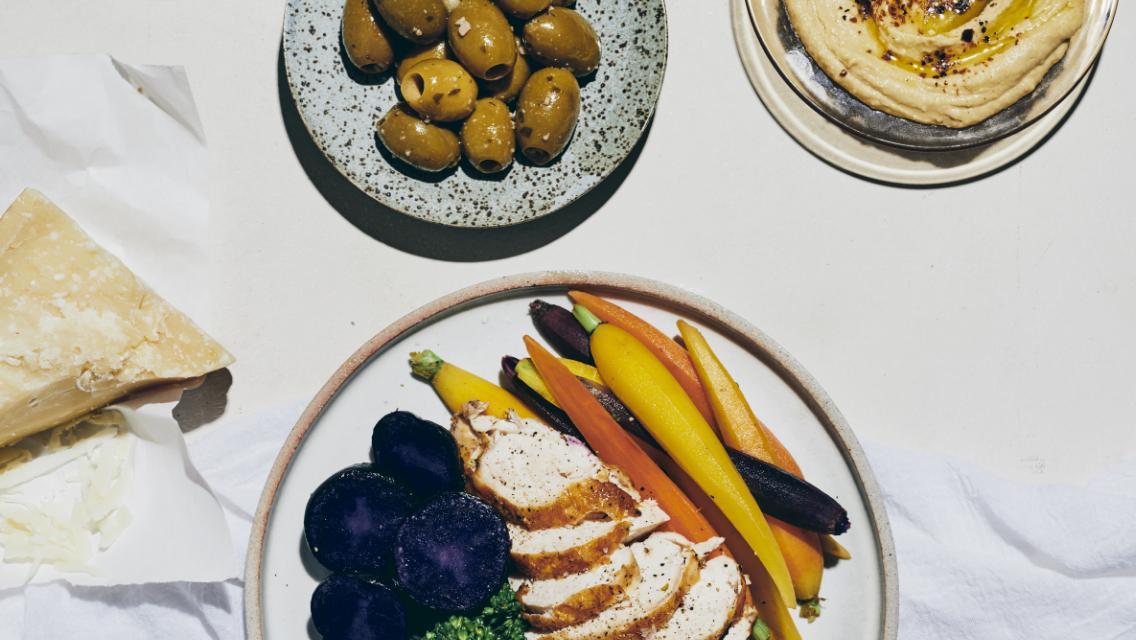 A spread of food on a table including a plate of purple potatoes, chicken breast, and carrots.