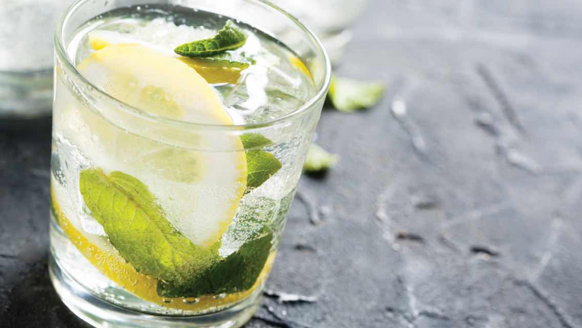 Clear drink with lemon and herbs in it on a black table.