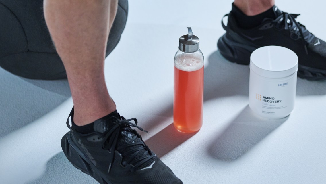 A person wearing black tennis shoes with a water bottle of pink liquid Amino Recovery