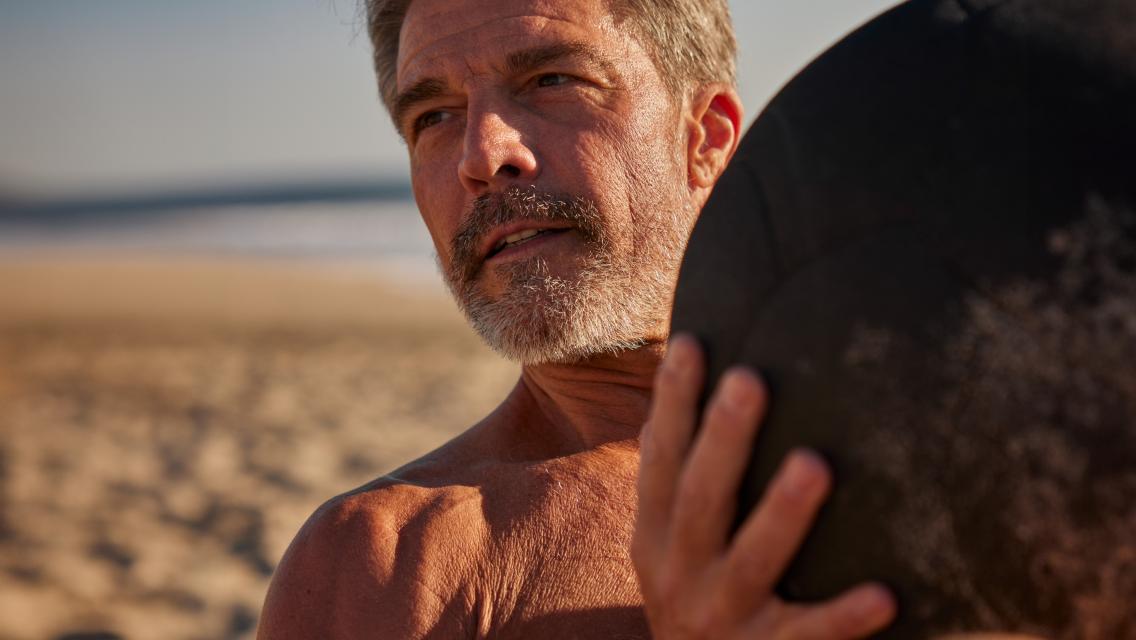 A male with gray hair exercising with a medicine ball on a beach.
