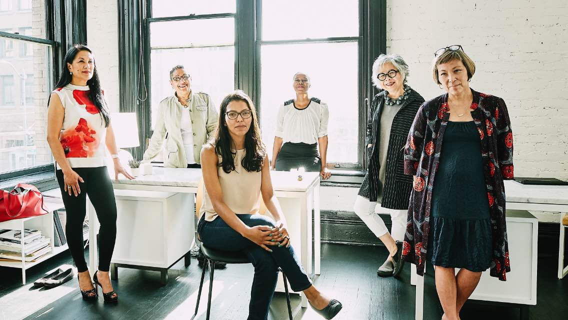 Seven women of various ages and ethnicities gathered in a work environment.