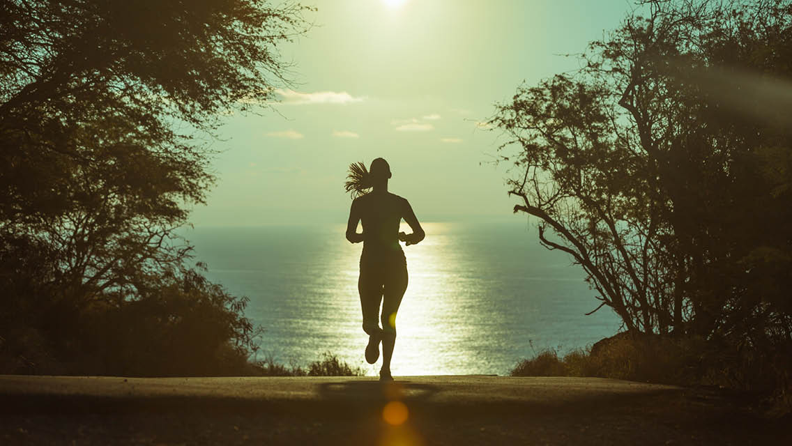 a woman runs through a shadowy woods towards a lake with the sun shining on it