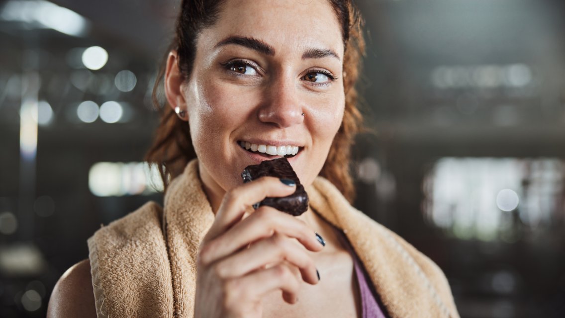 A woman eating a protein bar after a workout while in a health club.