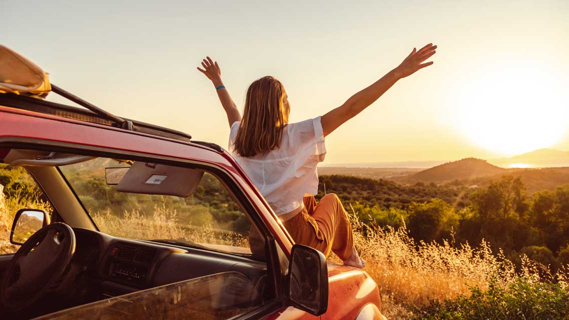 a women sits on the hood of her car with her arms extended at sunset